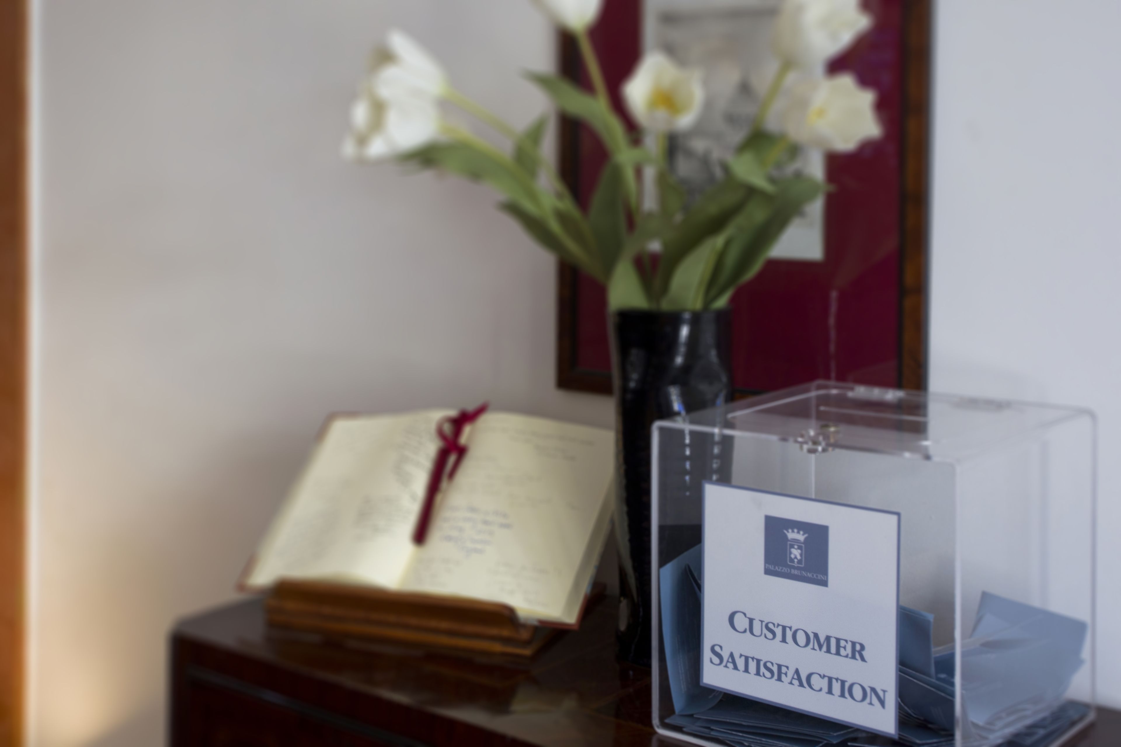 Una scatola di plastica trasparente con l'etichetta 'Customer Satisfaction', un libro e un vaso di fiori su un piano di legno.