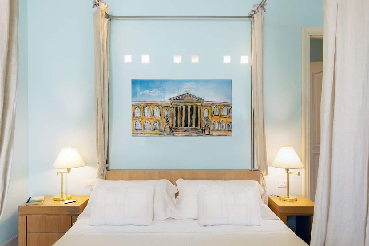Une chambre d’hôtel avec un lit, deux lampes de chevet, un tableau représentant un bâtiment classique et un rideau.