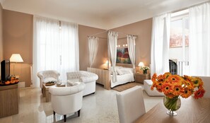 Habitación de hotel con cama con dosel, dos sillones, una mesita de café, un televisor y un jarrón con flores.