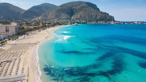A coastal beach with rows of sun loungers and umbrellas, a clear blue sea, and a mountainous backdrop. - Palazzo Brunaccini Boutique Hotel (Palermo)
