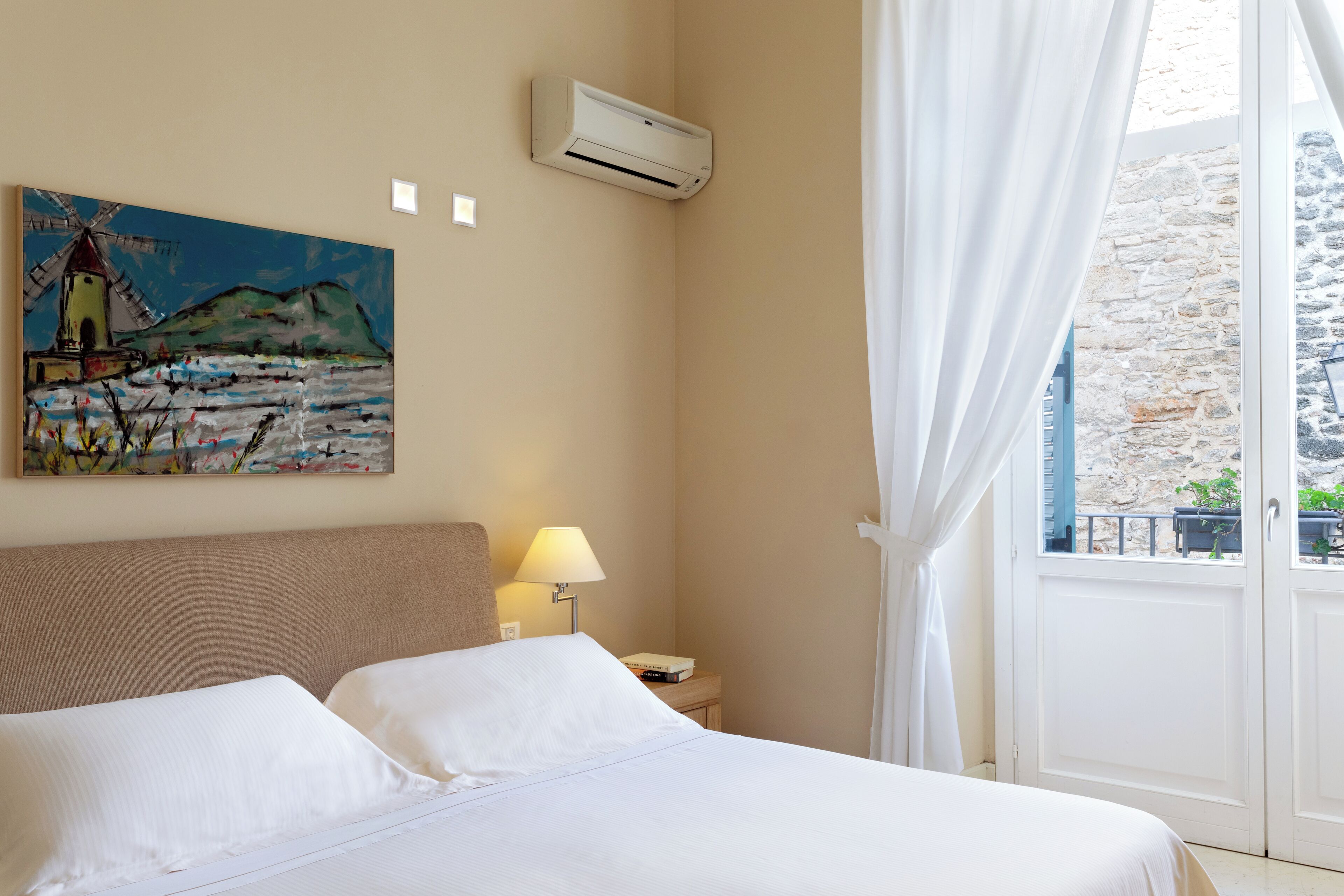 Habitación de hotel con cama, un cuadro en la pared, equipo de aire acondicionado, lámpara y vista a un edificio de piedra por una puerta.