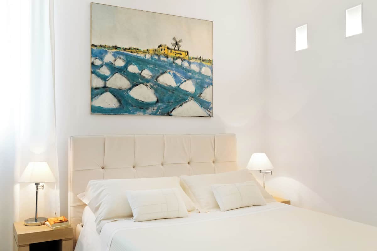 Une chambre à coucher avec un grand lit, deux tables de chevet avec des lampes, une tête de lit blanche et un tableau représentant un pont accroché au mur.