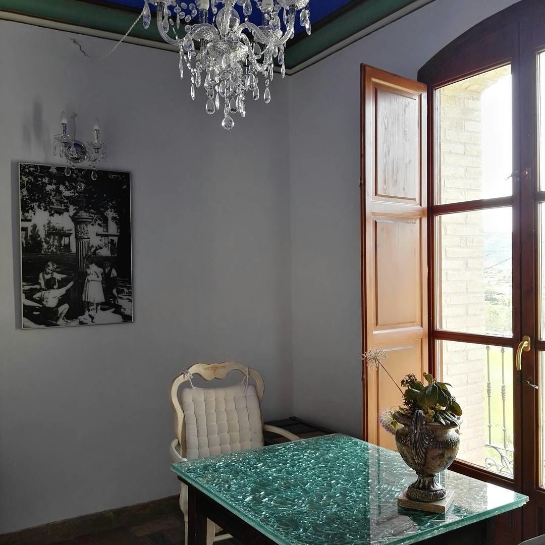 A room with a green marble table, a white upholstered chair, a chandelier, a black and white photograph on the wall, and a window with a view of a building.