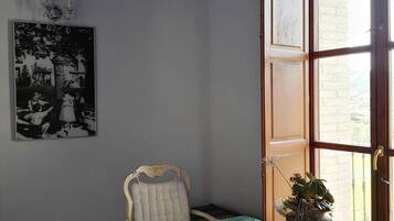 A room with a green marble table, a white upholstered chair, a chandelier, a black and white photograph on the wall, and a window with a view of a building.