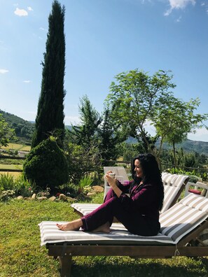 Una persona sentada en una silla reclinable a rayas al aire libre, con vistas a un paisaje montañoso y un árbol alto al fondo.
