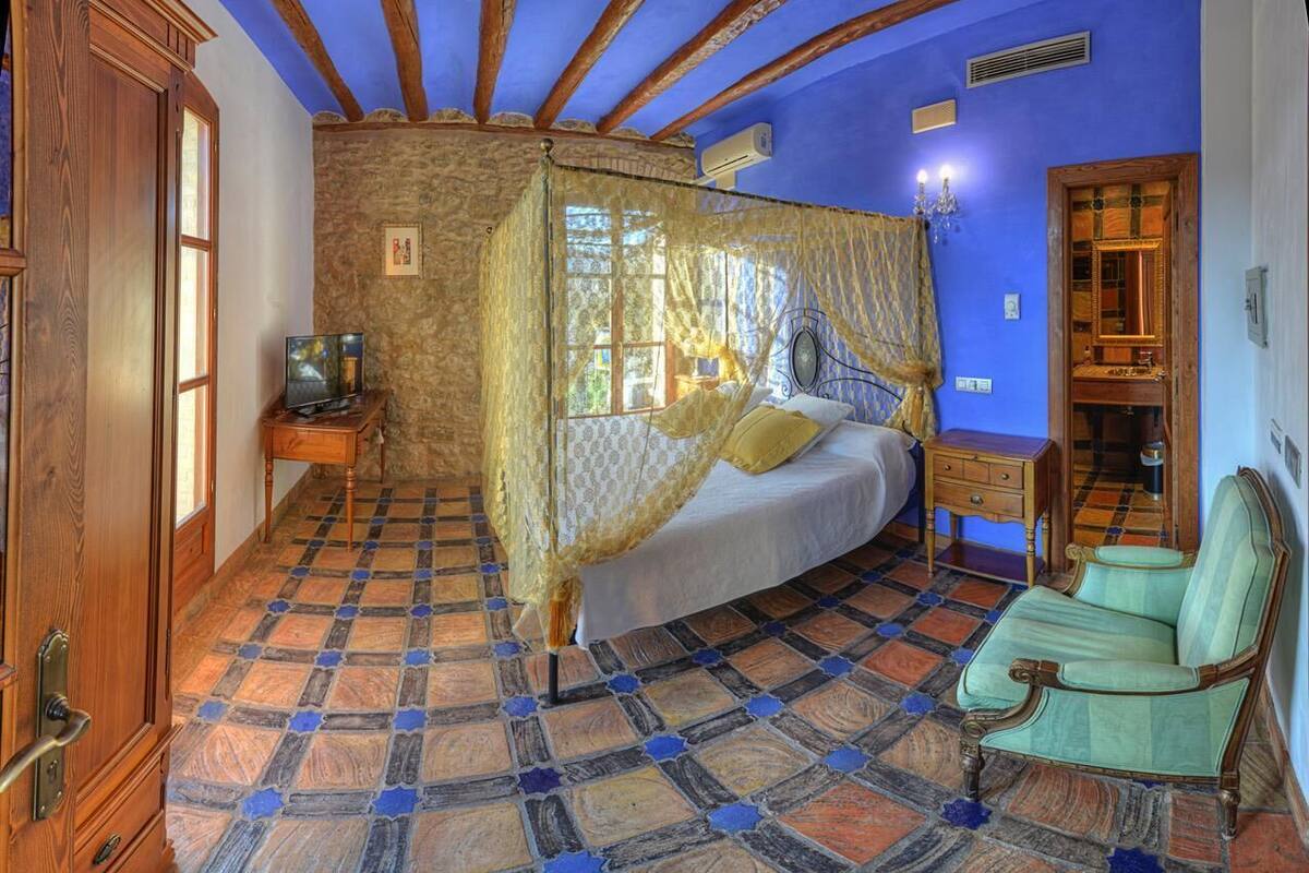A bedroom with a bed under a canopy, a wooden nightstand, a green armchair, and a stone wall.