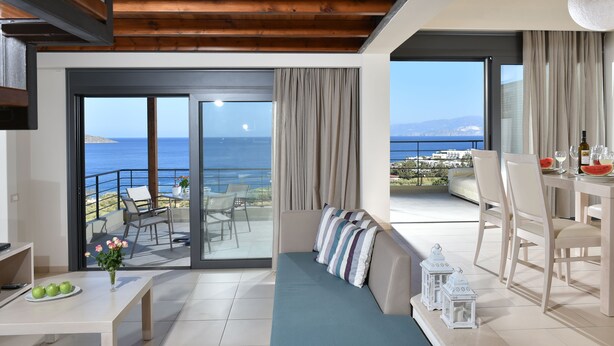 Un salon moderne avec un canapé, une table à manger et des chaises. Un balcon avec vue sur la mer est visible à travers de grandes portes vitrées.