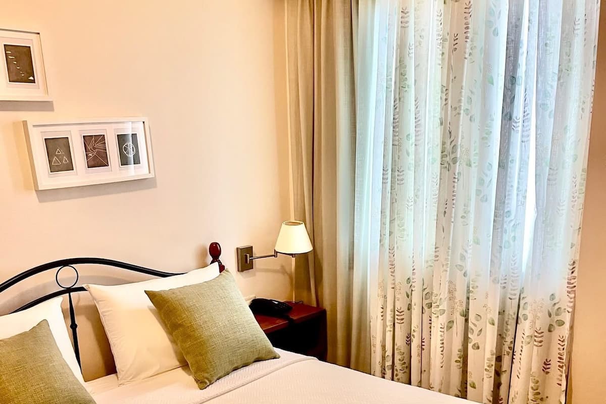 Een net opgemaakt bed met een beddekleed met patroon en kussens, een bedlampje en een raam met bloemen gordijnen.