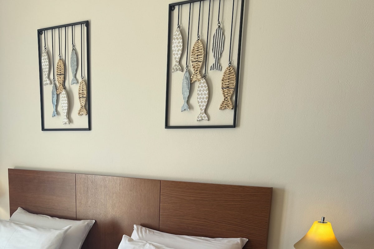 Een net opgemaakt bed met witte lakens en een dekenovertrek met een herringbone-patroon, een houten hoofdbord, twee vis kunstwerken aan de muur, een bedlampje en een nachtkastje met een lamp en een glas.