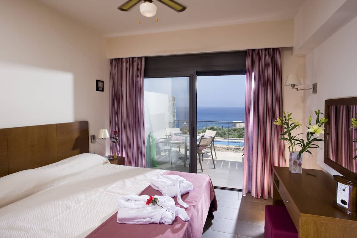 Hotelkamer met een bed, bureau, stoel, een vaas met bloemen en uitzicht op de oceaan door een schuifdeur.