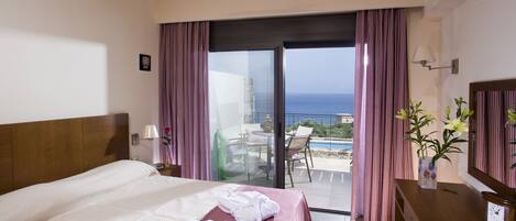 A hotel room with a bed, a desk, a chair, a vase with flowers, and a view of the ocean through a sliding door.