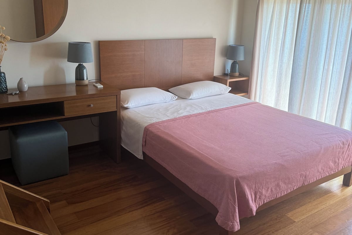 Een hotelkamer met een houten bed, een bureau met stoel, een klein tafeltje met lamp, een spiegel en een raam met gordijnen.