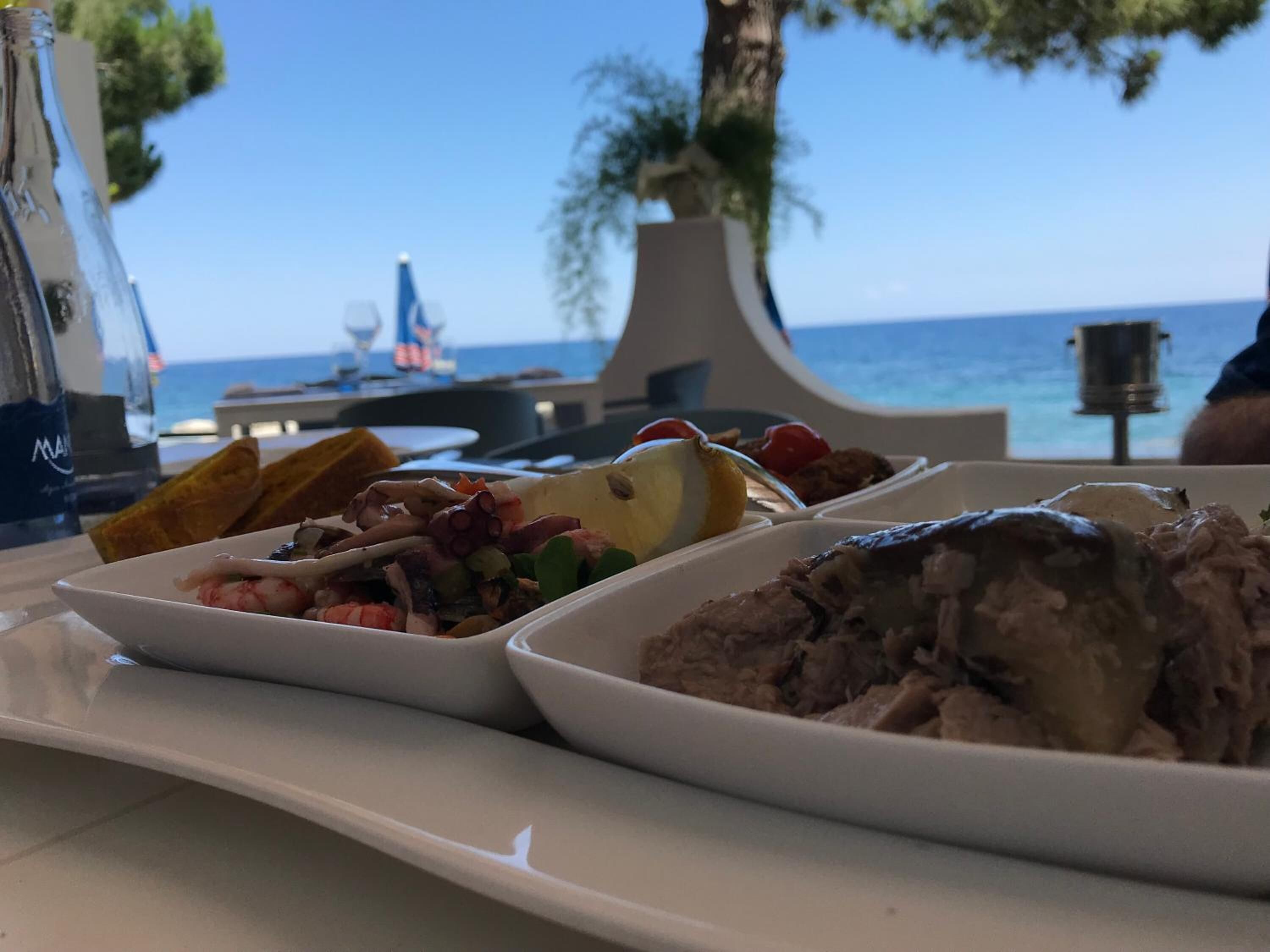 A dining table with seafood dishes and bread, set against a backdrop of a clear blue sky and ocean.