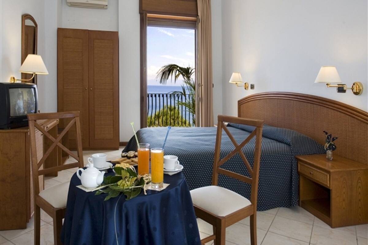 A hotel room with a bed, a table with breakfast items, a TV, and a view of the sea.