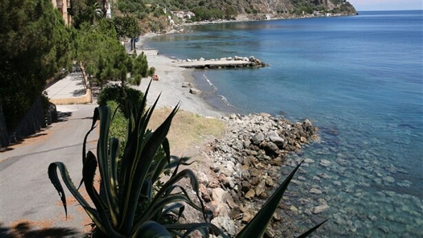 A coastal road with a rocky shoreline, clear water, and a hillside with buildings. - Hotel Riviera Lido (Milazzo)
