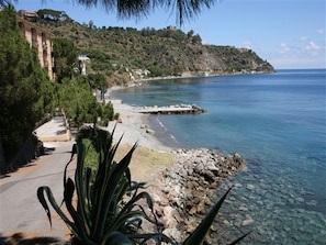 A coastal road with a rocky shoreline, clear water, and a hillside with buildings. - Hotel Riviera Lido (Milazzo)