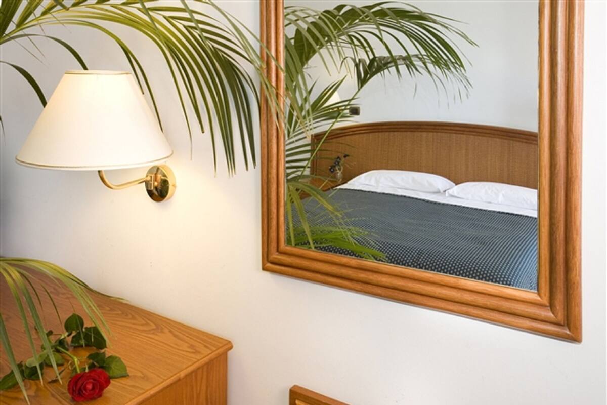 A hotel room with a wooden headboard, a bedside lamp, a mirror reflecting the bed, and a wooden nightstand with a red rose.