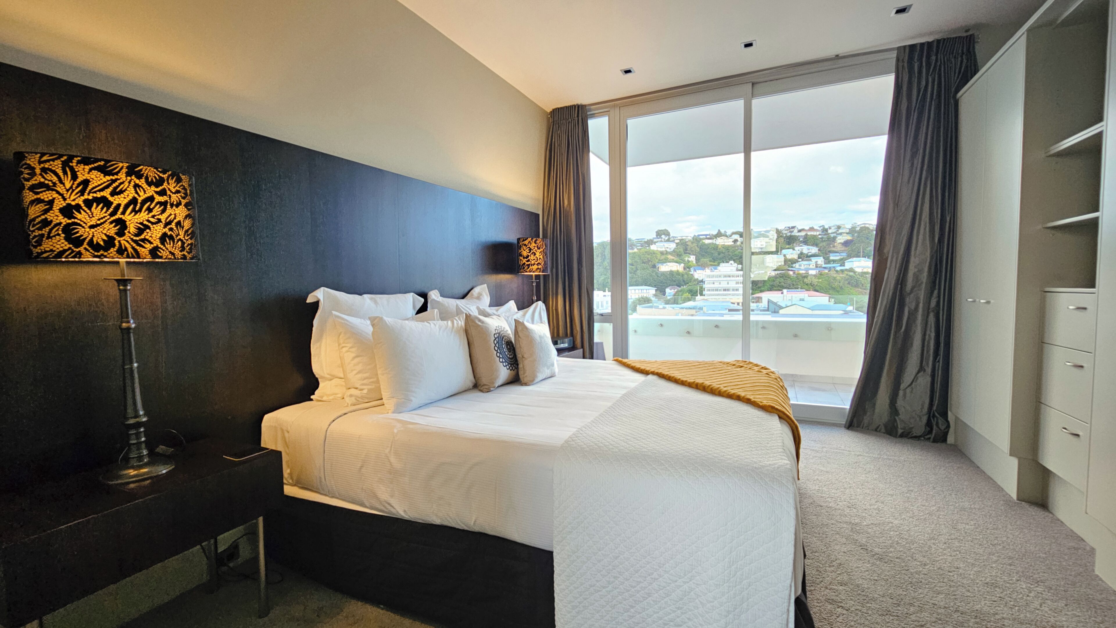 Habitación de hotel moderna con una cama grande, una mesita de noche, una lámpara y vistas a la ciudad a través de amplios ventanales.