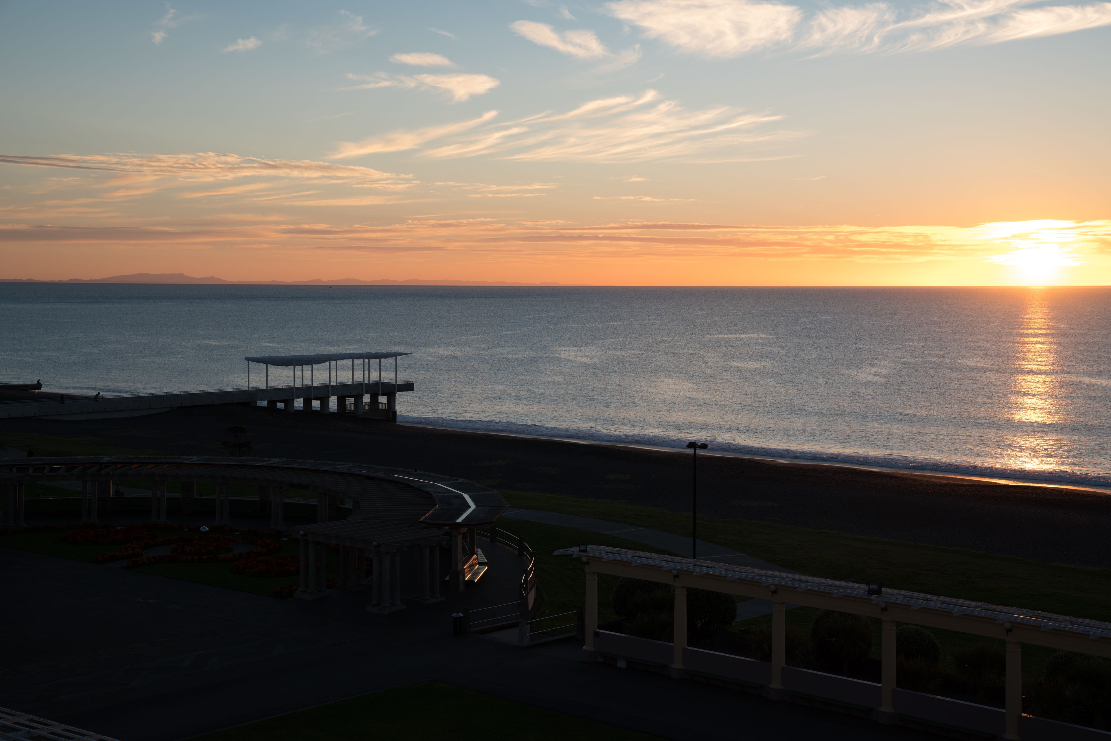 Vista costera con un atardecer sobre el agua, un muelle y un pequeño edificio con techo.