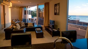 A modern living room with a sofa set, a coffee table, and a TV. There is a balcony with a view of the sea.