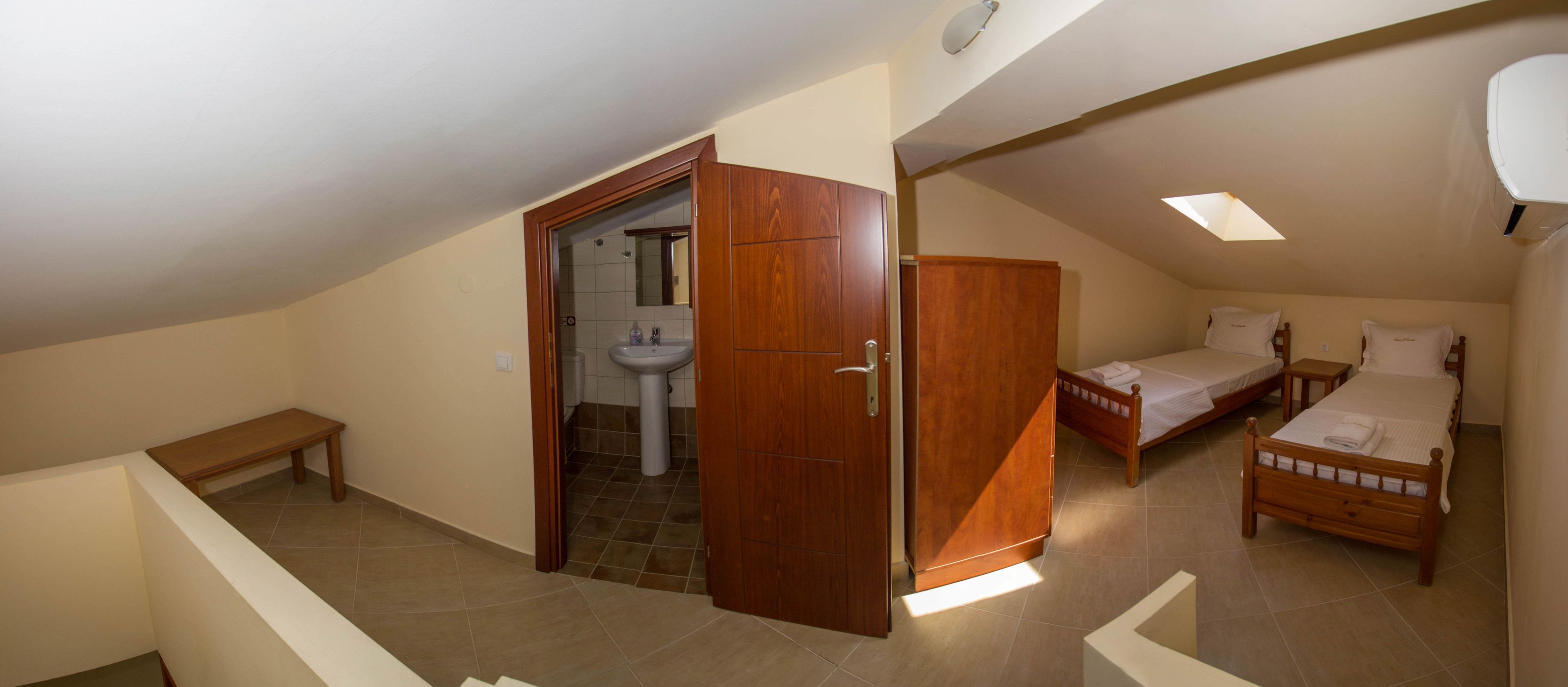 A bedroom with two wooden beds, a wooden wardrobe, a bathroom with a sink and mirror, and a small bench.