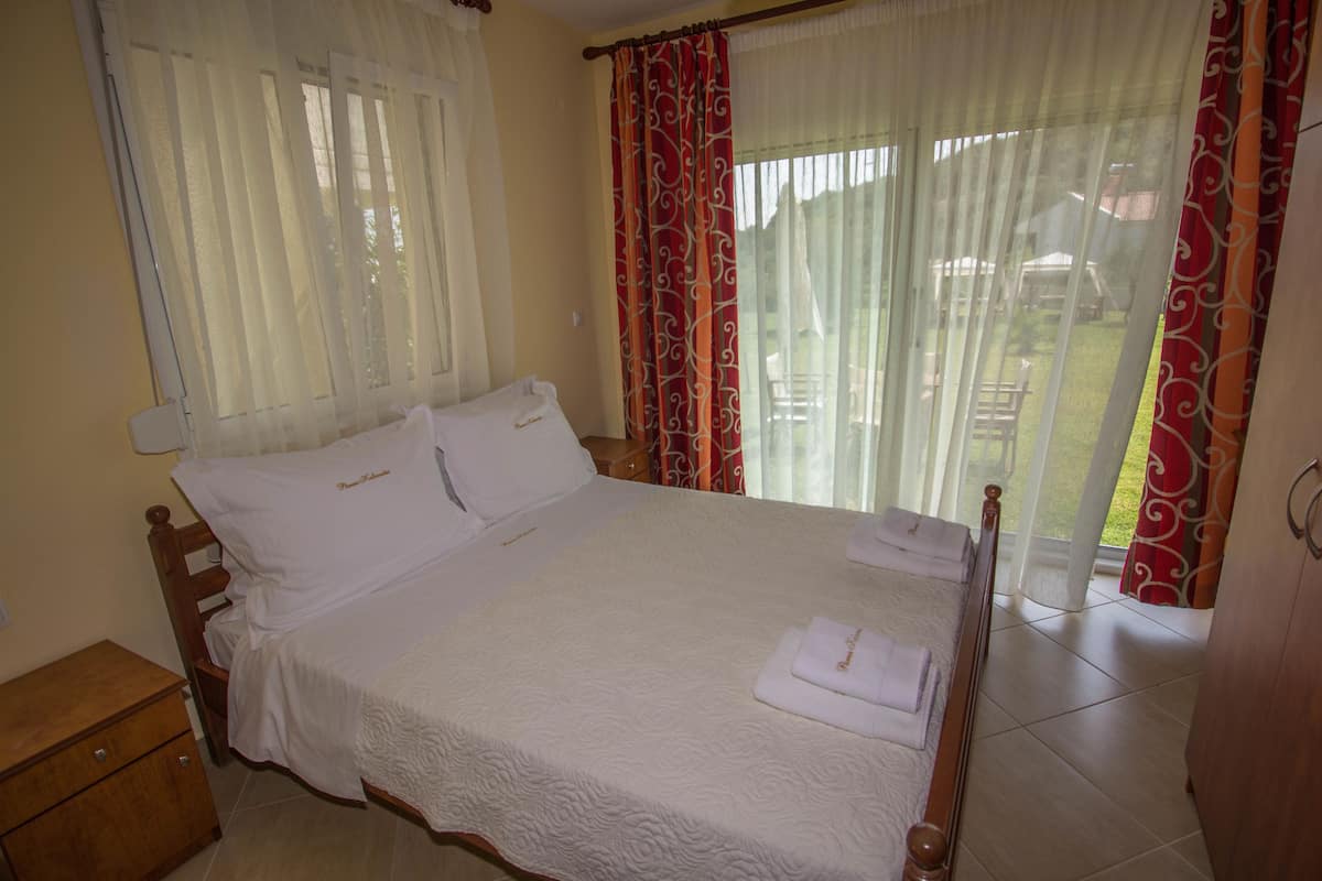 A neatly made bed with white linens and a wooden headboard, a wooden nightstand with drawers, and a view of a garden through sheer curtains.