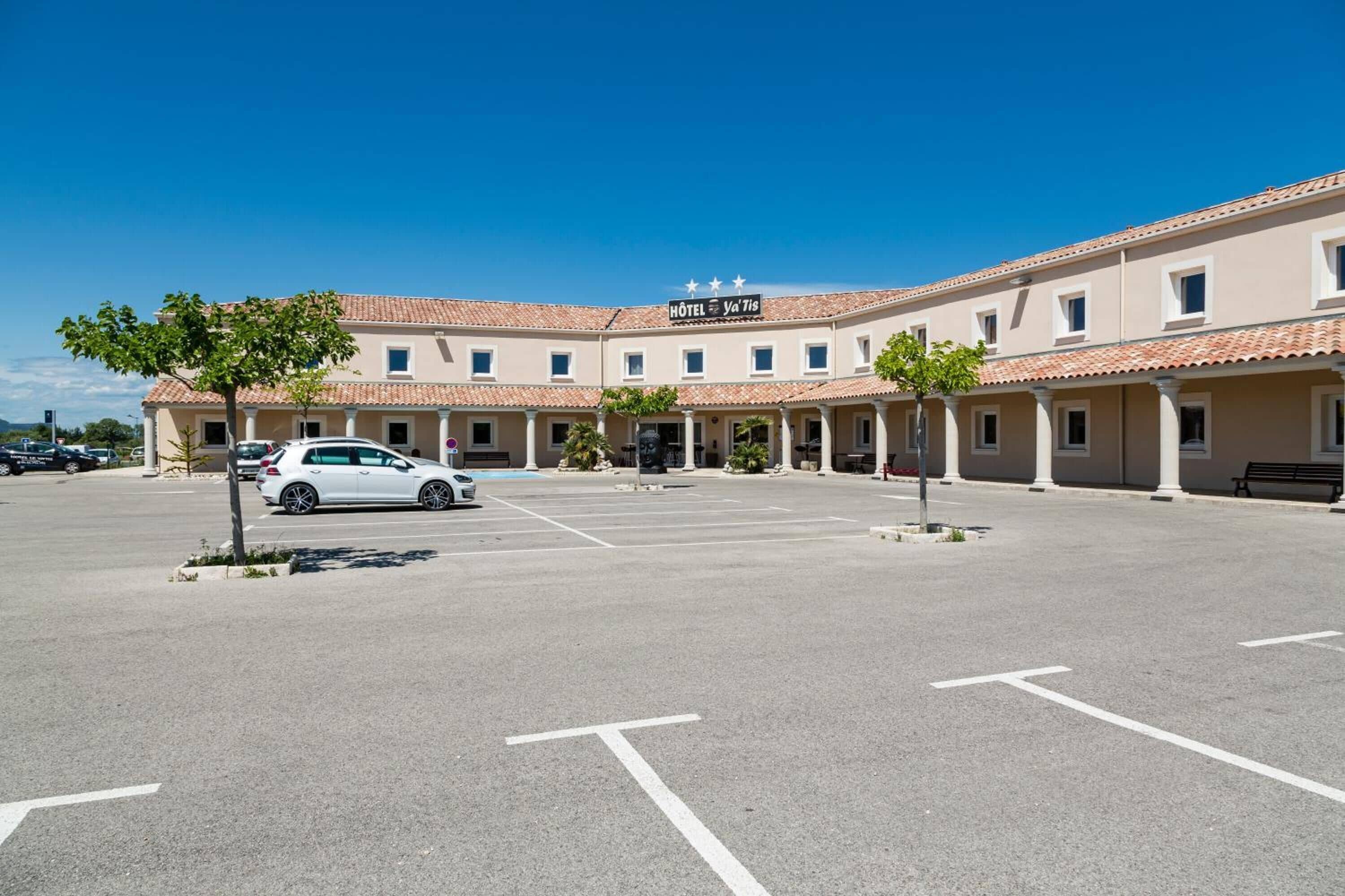 A hotel with a tiled roof, a sign reading "HOTEL de la Tige," and a parking lot with marked spaces.