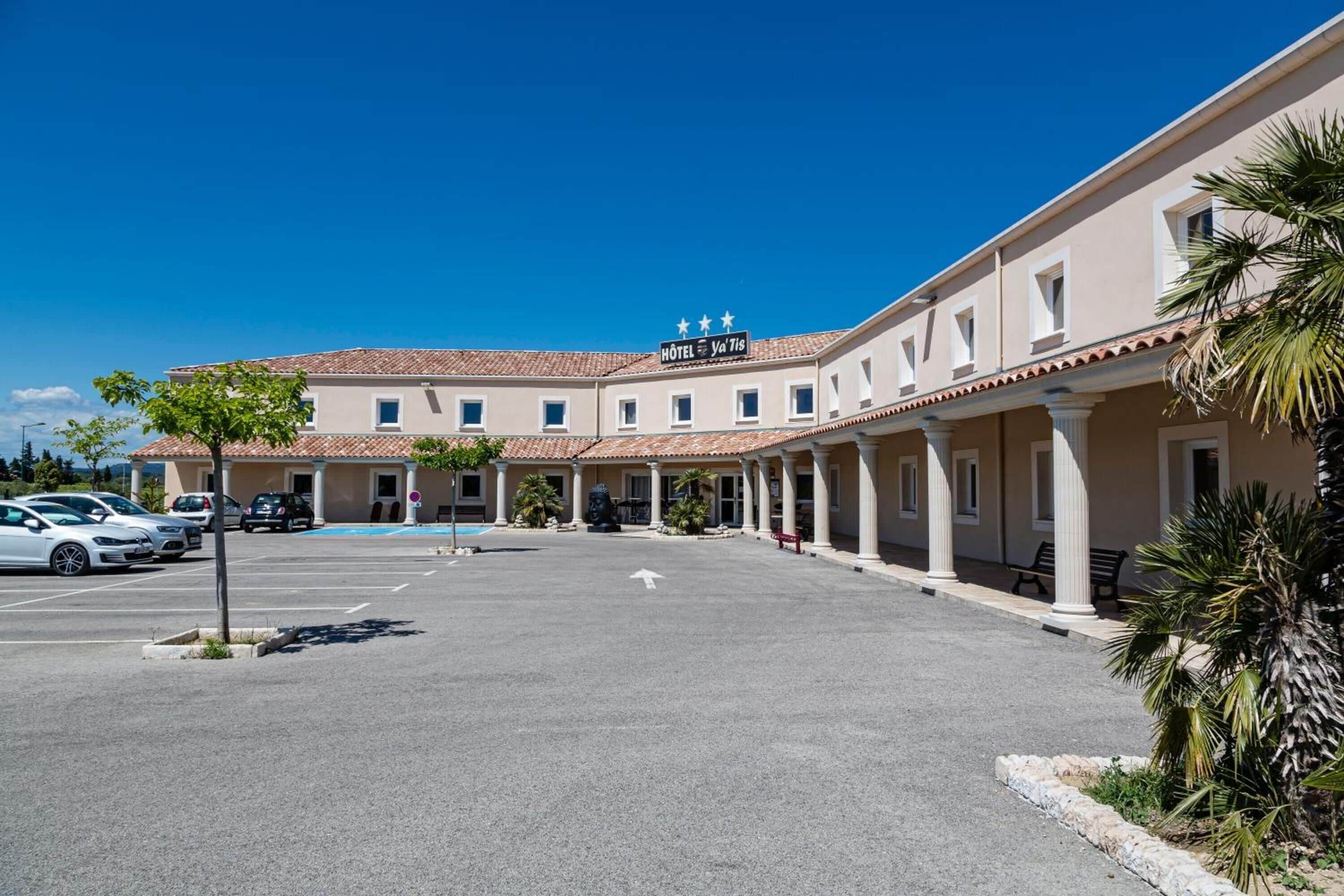 A hotel with a central courtyard, a sign reading "HOTEL Va Tis," and a parking lot with several cars.