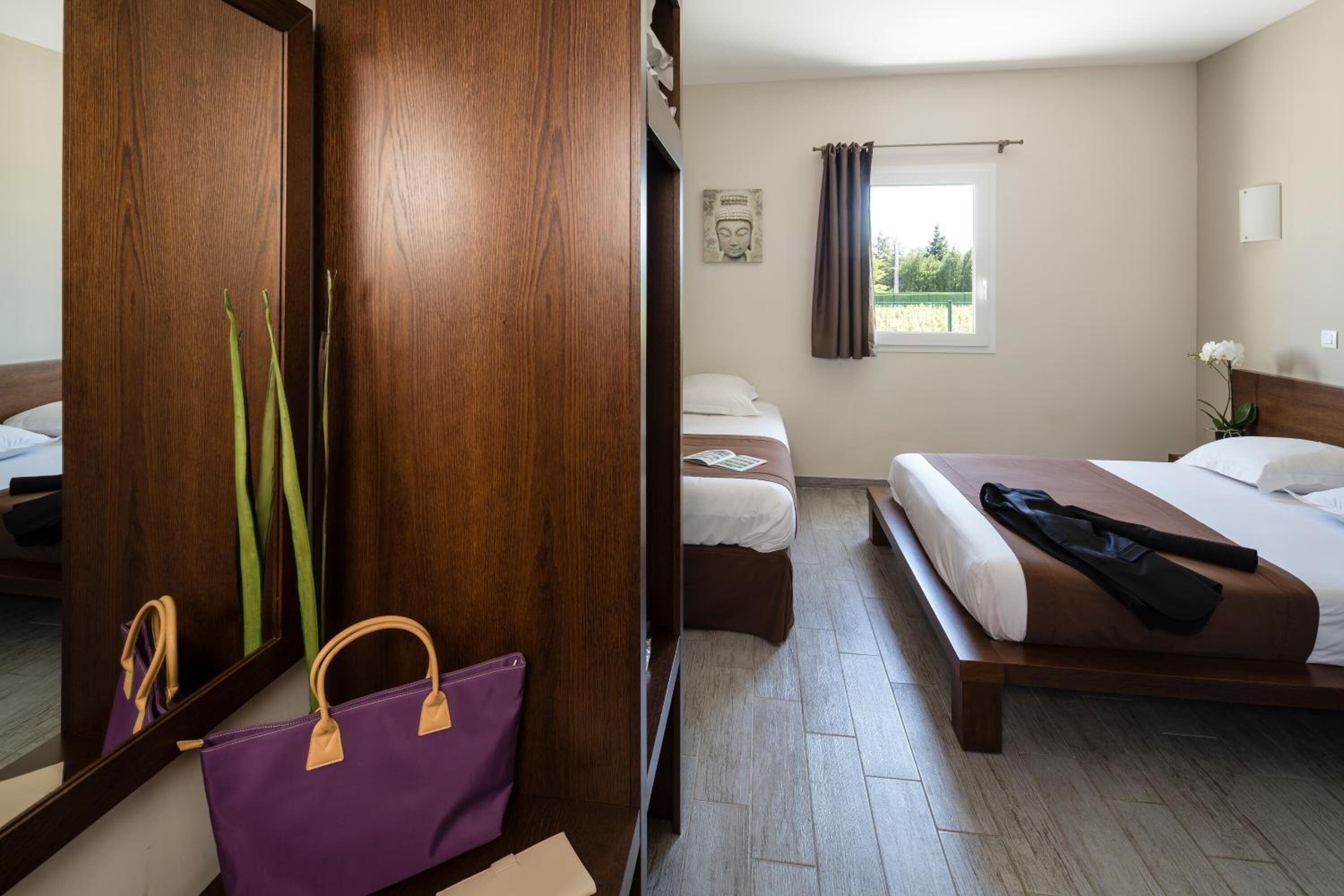 A hotel room with a wooden wardrobe, a bed with a brown headboard, a bedside table, and a window with curtains.