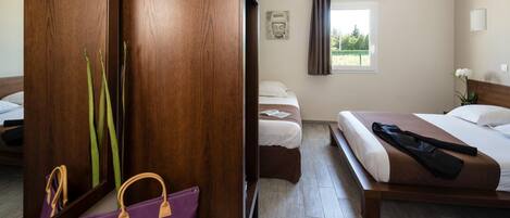 A hotel room with a wooden wardrobe, a bed with a brown headboard, a bedside table, and a window with curtains.