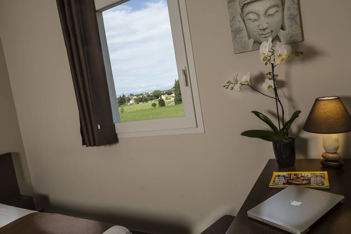 Een hotelkamer met een bed, een bureau met een laptop, een lamp, een potorkidee, een Boeddha-schilderij en een raam met uitzicht op een groen landschap.
