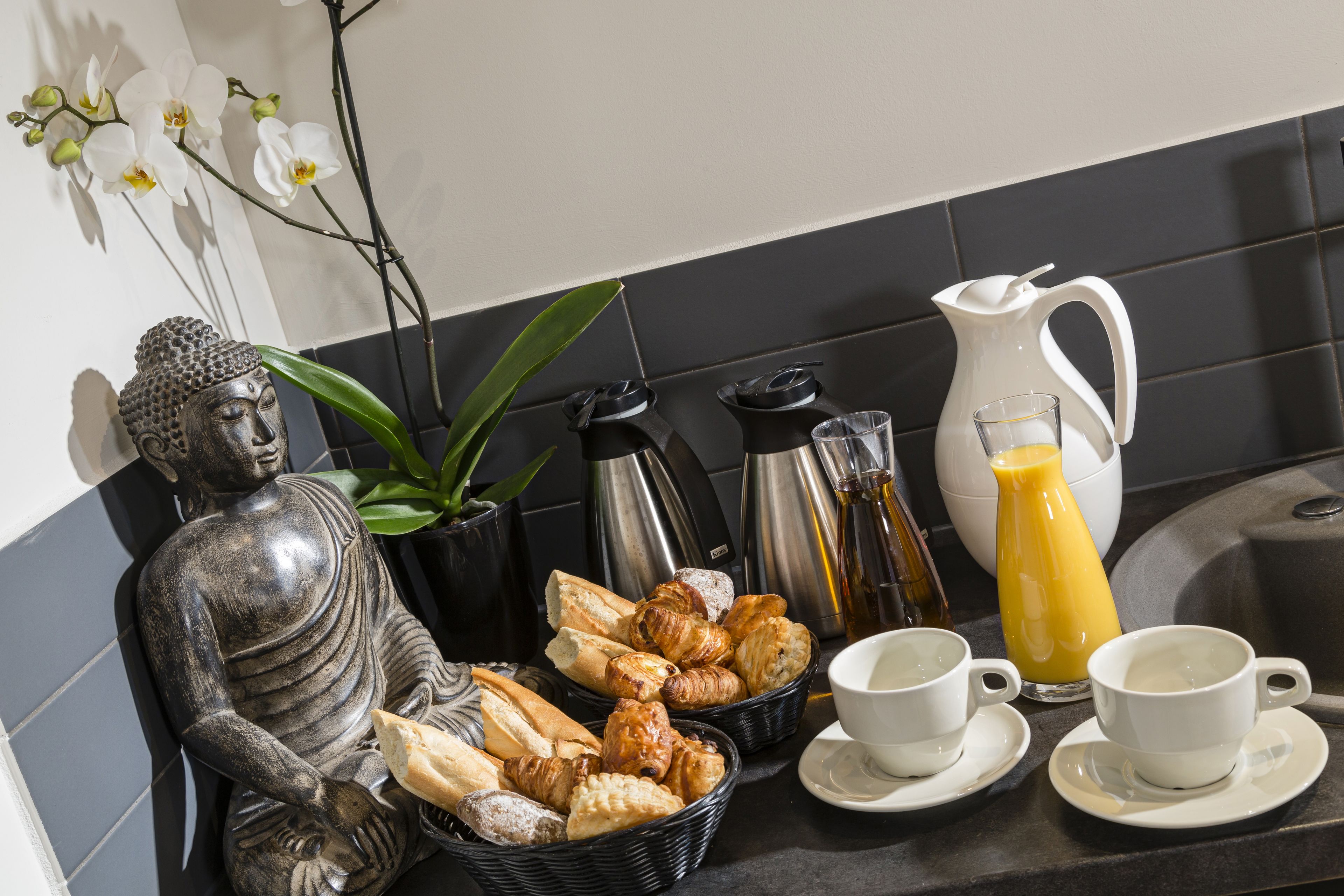 A breakfast setup with croissants, coffee, and juice, accompanied by a Buddha statue and orchids.