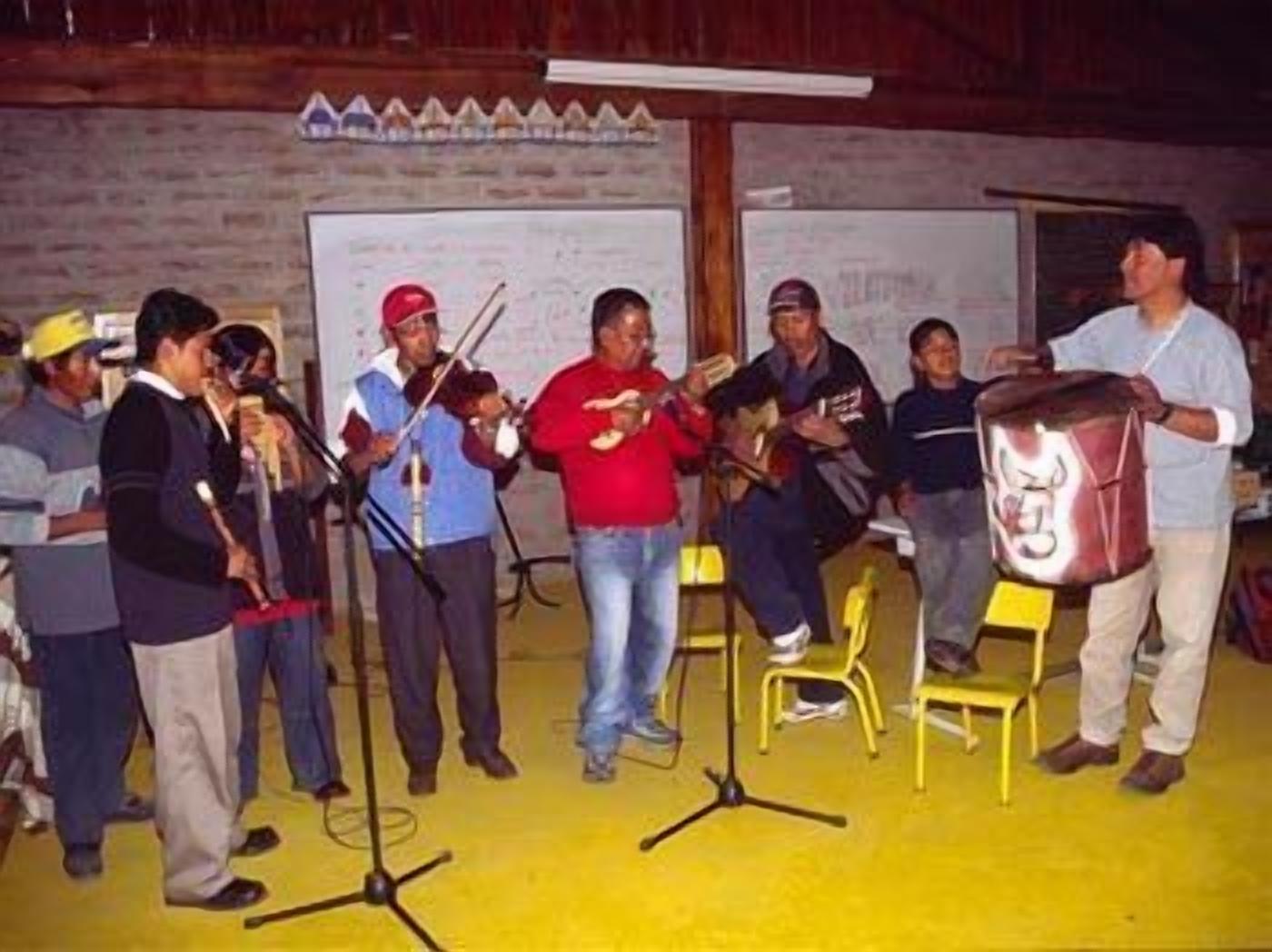 Um grupo de pessoas se apresentando em um palco com instrumentos musicais, em uma sala com piso amarelo e paredes de tijolo.