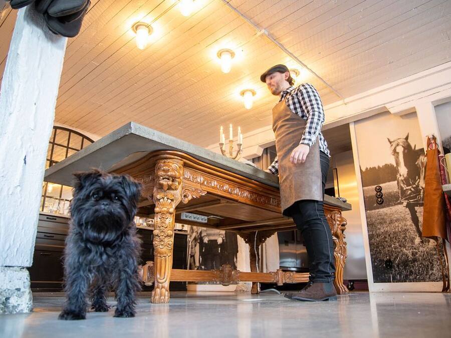 Una persona de pie junto a una mesa de madera con adornos labrados, un perro pequeño y un cuadro en la pared.