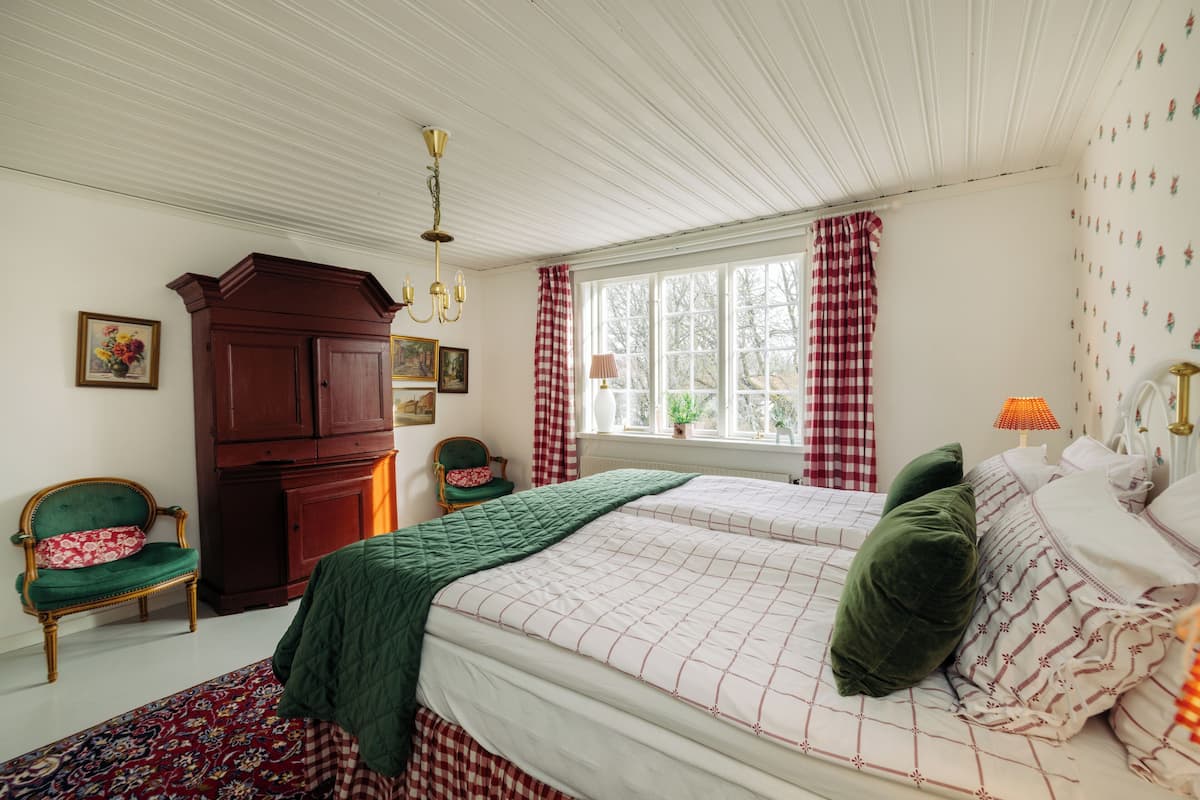 Ein Schlafzimmer mit zwei Betten, einem Holzschrank, einem Sessel und einem Fenster mit Vorhängen.