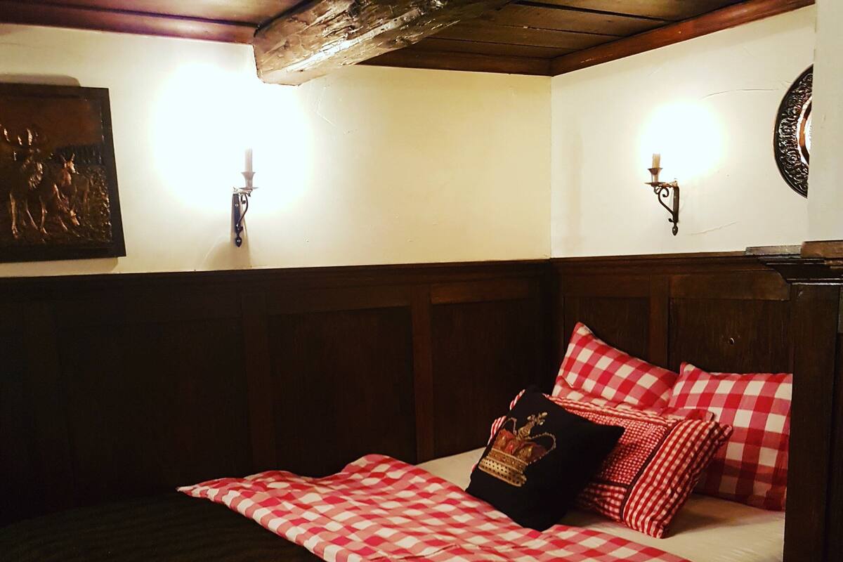 Ein Bett mit karierter Decke, einem Kopfkissen mit einem Krondruck und einer Wandleuchte.