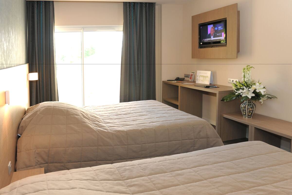 Habitación de hotel con dos camas, un televisor montado en la pared, un escritorio con un jarrón de flores y una ventana con cortinas.