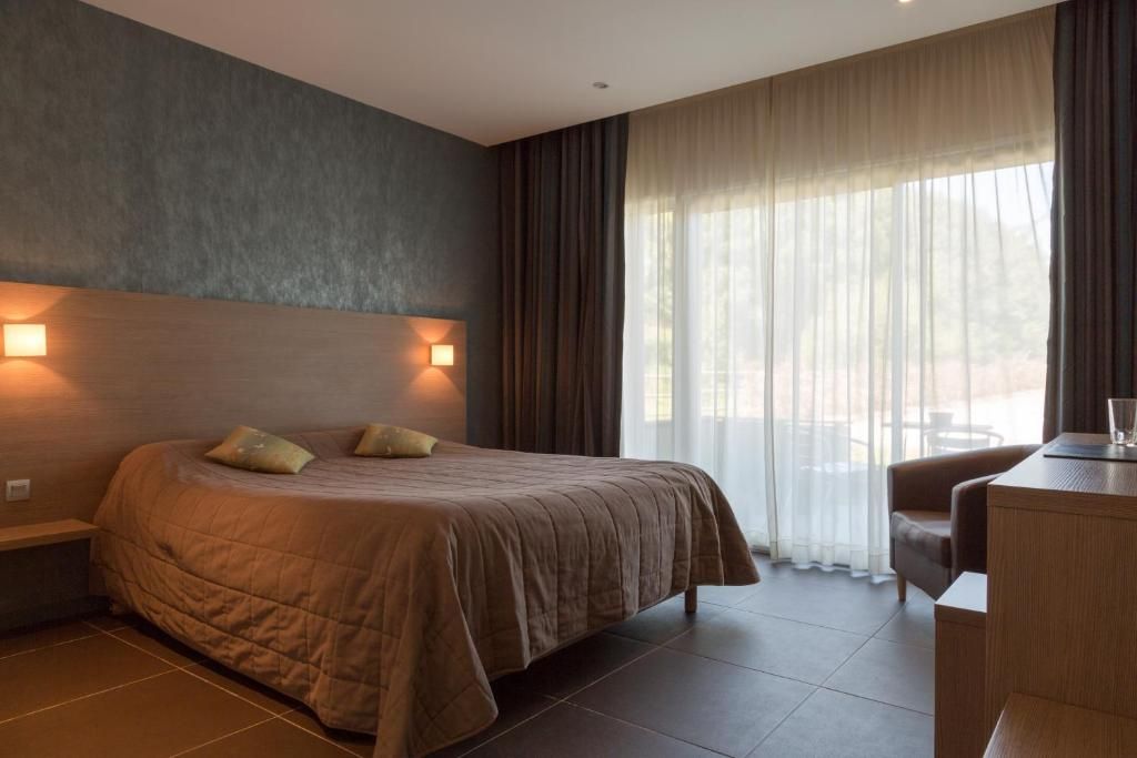 A modern hotel room with a large bed, a desk, and a view of the outdoors through sheer curtains.