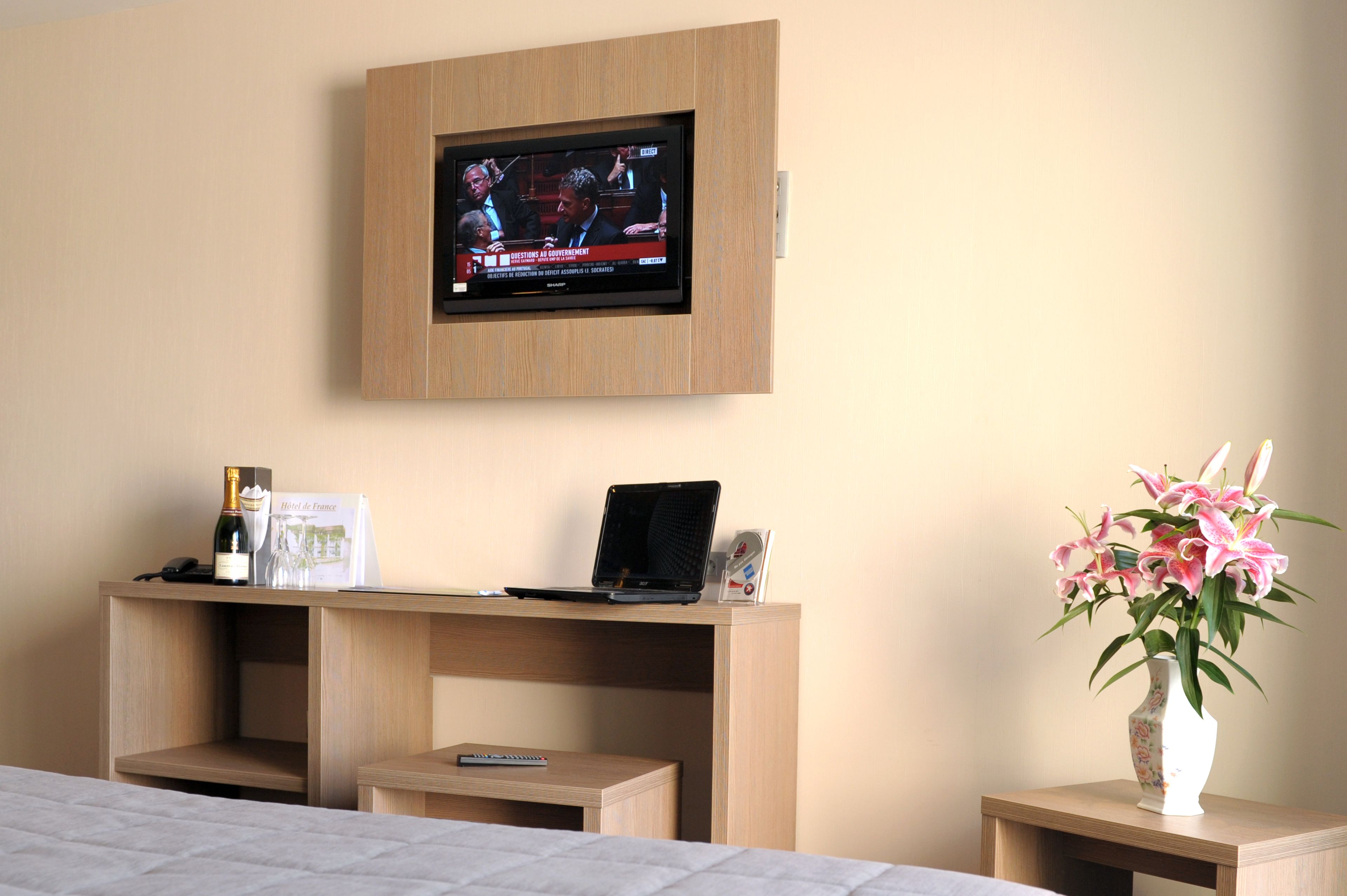 A hotel room with a TV mounted on the wall, a laptop on a wooden desk, a remote control, a bottle of wine, a vase with flowers, and a bed with a quilted cover.