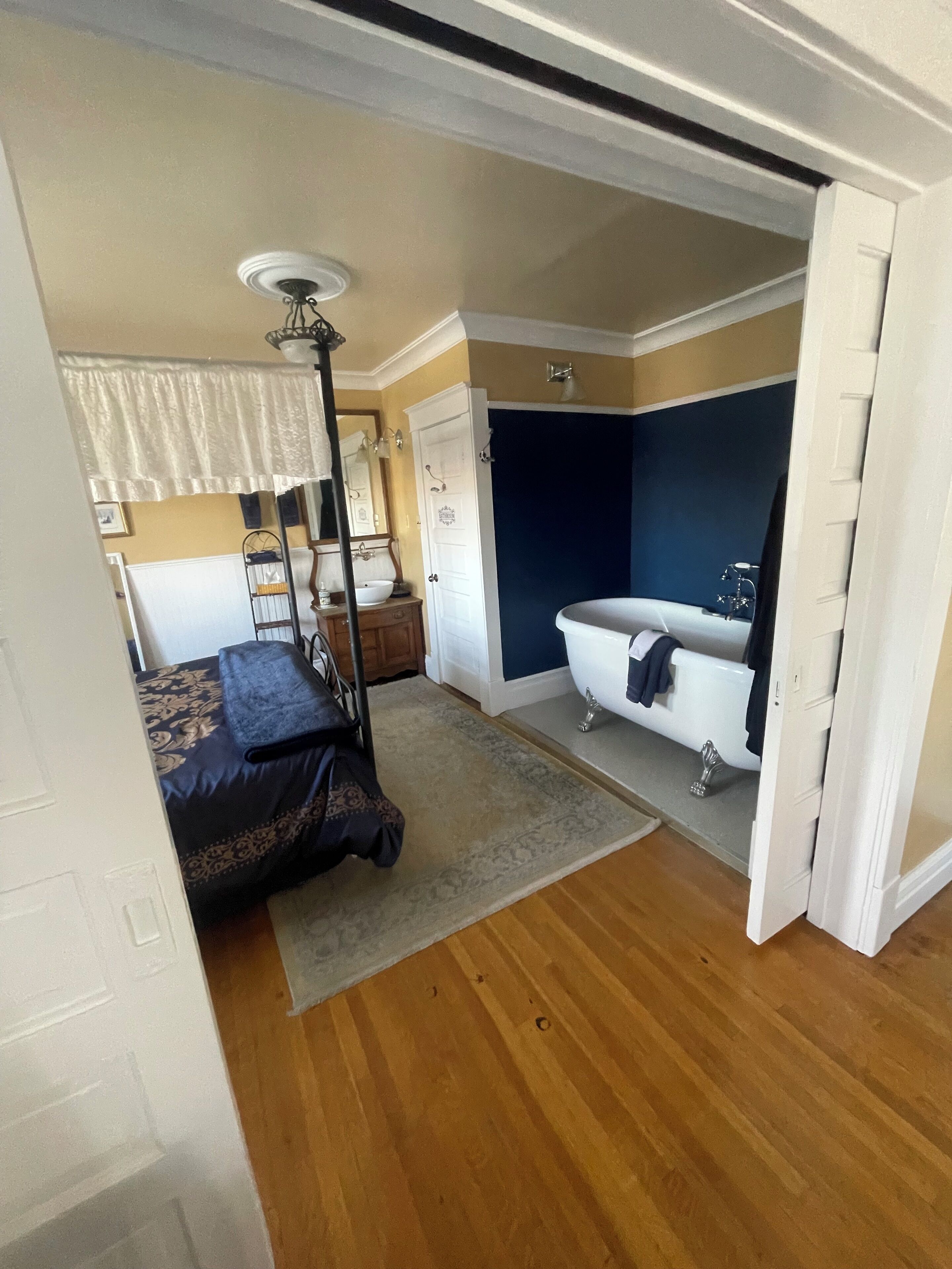 A compact bedroom with a single bed, a small bedside table, a nightstand, a dresser, a vanity with a mirror, a ceiling fan, and a bathtub.