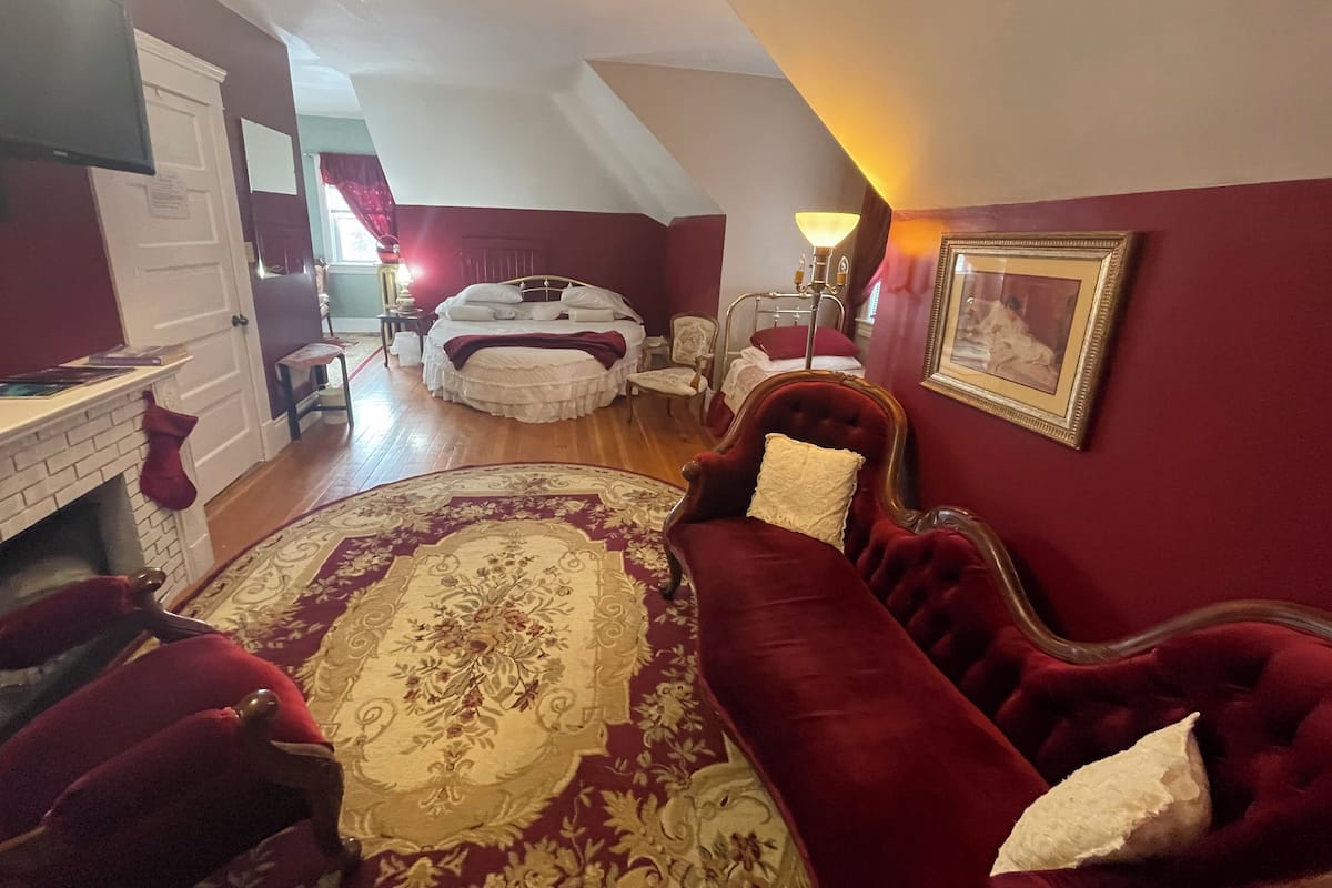 A bedroom with a large bed, a red velvet sofa, a wooden desk, and a fireplace.