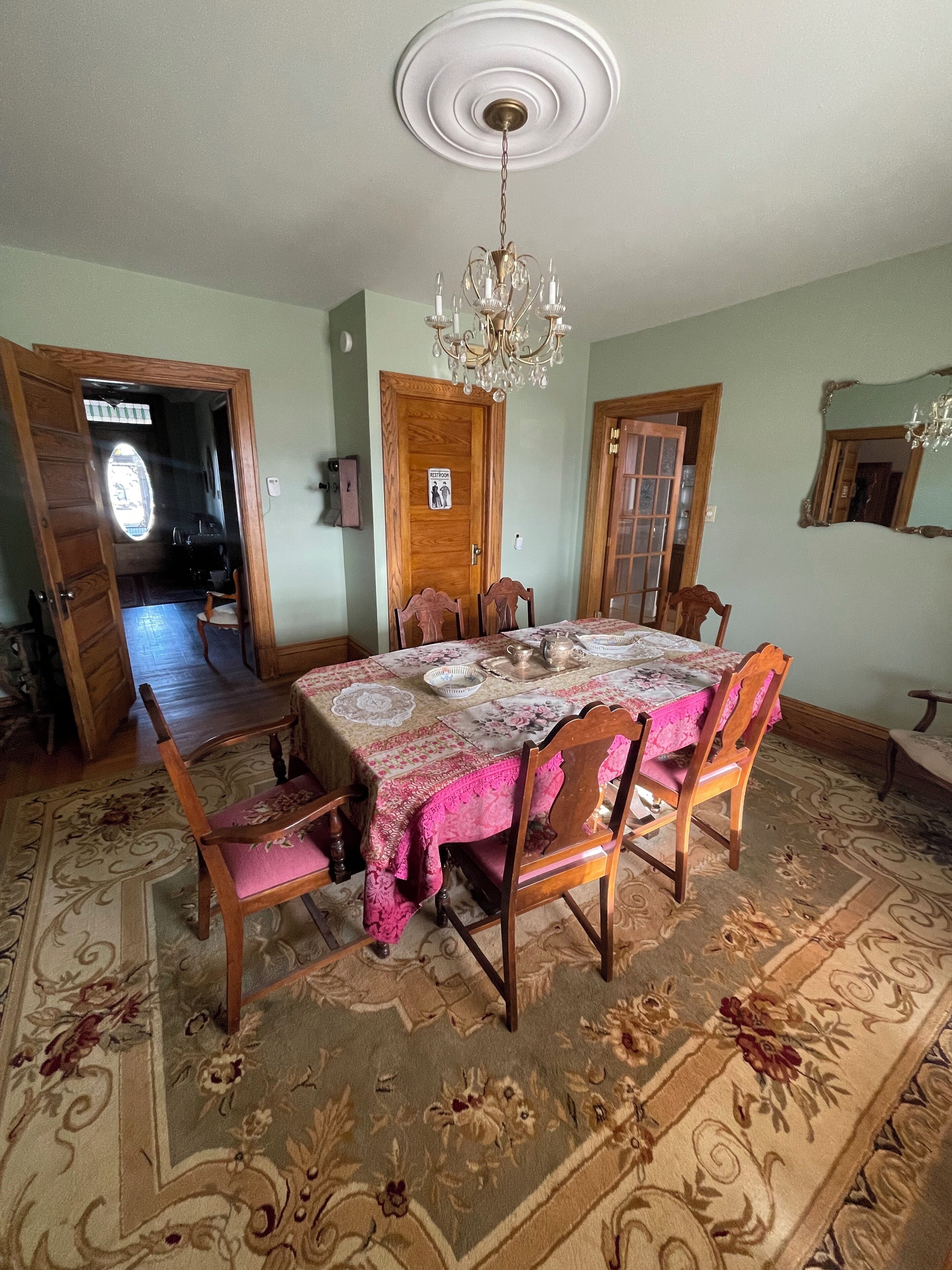 Une salle à manger avec une table dressée pour quatre personnes, entourée de chaises en bois. Un lustre est suspendu au plafond et la pièce est agrémentée d’un tapis à motifs.