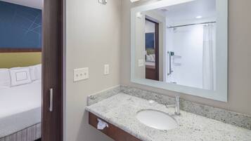 A hotel bathroom with a granite countertop sink, a large mirror, and a shower visible in the reflection.