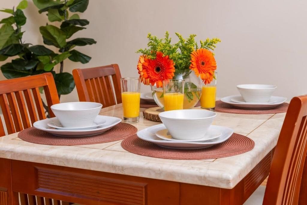 Een eettafel gedekt met witte kommen, borden en glazen, met jus d'orange en een vaas oranje bloemen.
