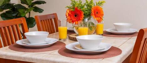 Een eettafel gedekt met witte kommen, borden en glazen, met jus d'orange en een vaas oranje bloemen.