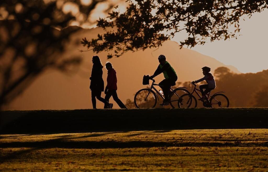 Silhouettes di persone che camminano e vanno in bicicletta in un parco, con una montagna sullo sfondo.
