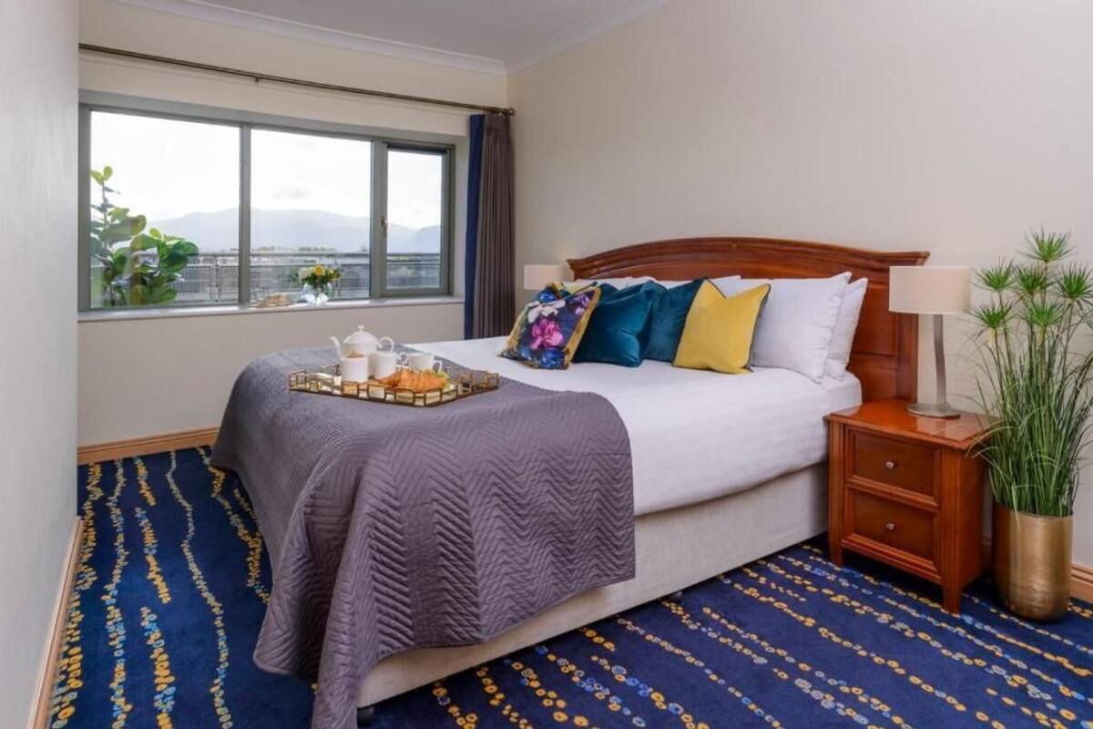 A hotel room with a bed, a window with a view, a bedside table with a lamp, and a decorative plant.