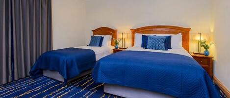 A hotel room with two beds, each with a blue bedspread and matching pillows. There are bedside tables with lamps and a vase of flowers. The room has a blue carpet with a pattern.