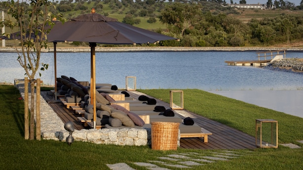 Een rustig buitenzitgedeelte met loungestoelen en een zonnescherm, met uitzicht op een meer met een steiger en een grasrijk landschap op de achtergrond.