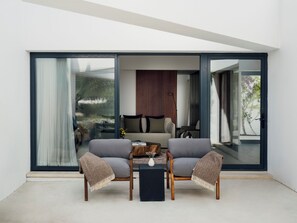 A modern living room with a sofa, two armchairs, a coffee table, and a large sliding glass door leading to a balcony with a view of greenery.